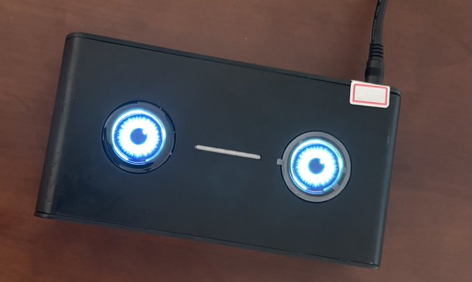 The image shows a sleek black rectangular device with two glowing blue circular "eyes" resembling a robotic face, connected by a power cable—the Helium Deploy Glod P3 miner, presented as a stylish desktop centerpiece rather than a typical industrial mining rig.