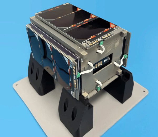 A CubeSat-style small satellite with black solar panels and a metallic body, mounted on a black display stand against a bright blue background.