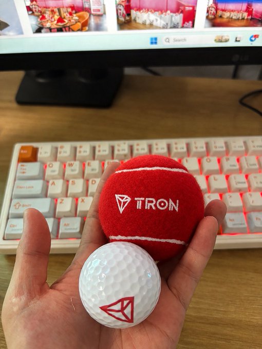 A hand holding TRON-branded merchandise including a red tennis ball and a white golf ball, with a mechanical keyboard and LG monitor visible in the background.