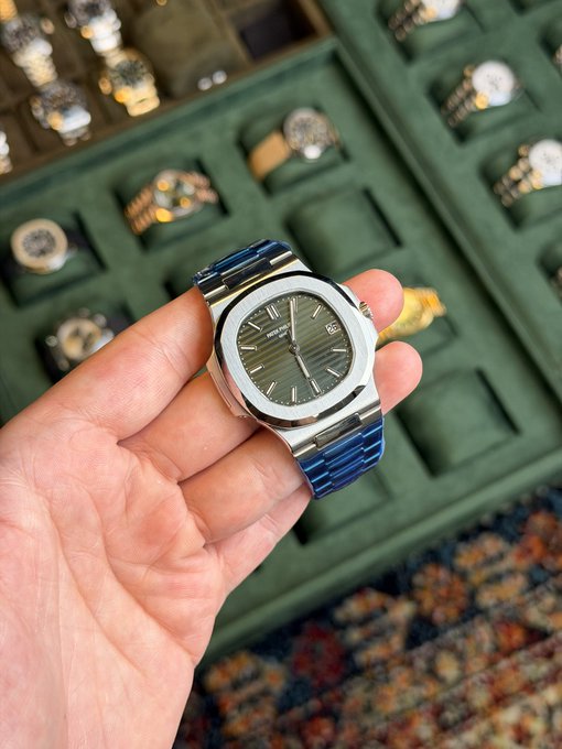 A hand holds a Patek Philippe Nautilus watch with a blue dial and steel bracelet, with other luxury watches visible in display cases in the background. The caption "Sold" indicates the watch has been successfully purchased or sold.