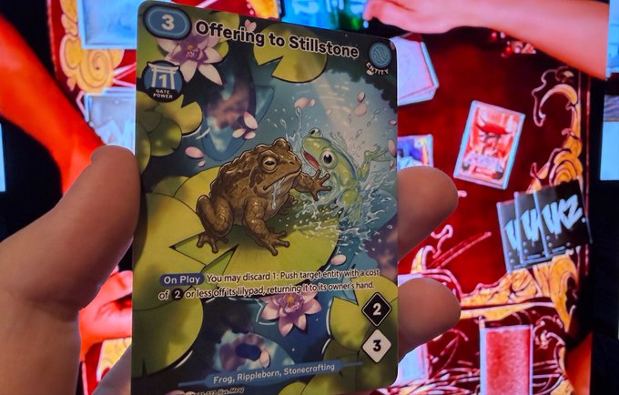 A hand holds up a trading card titled 'Offering to Stillstone' (cost 3), depicting a frog interacting with a water-like creature, with game text explaining its 'On Play' ability to push entities with a cost of 2 or less.