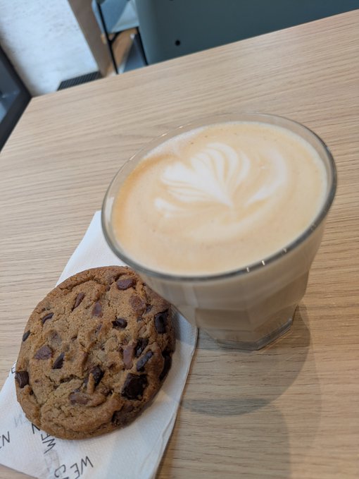 A latte with leaf-shaped latte art in a glass cup alongside a large chocolate chip cookie, served on a wooden table with a branded napkin.