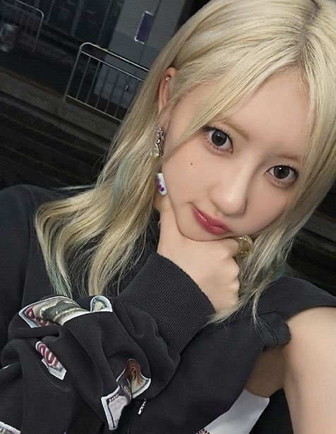 A close-up selfie of a young woman with blonde hair and dark roots wearing large earrings and a black top with decorative patches.
