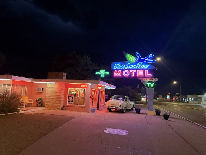 The illuminated neon sign of the 'Blue Swallow Motel' glows in pink, blue, and green at night, with a classic car parked in front on Route 66.