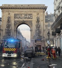 A street scene in Paris featuring an emergency response at the Arc de Triomphe area, with a fire truck, workers in orange uniforms, and debris visible on the road during what appears to be a protest or incident.