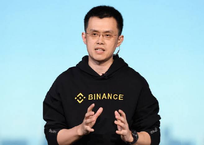 A man wearing glasses and a black hoodie with the "BINANCE" logo, gesturing with his hands while speaking.