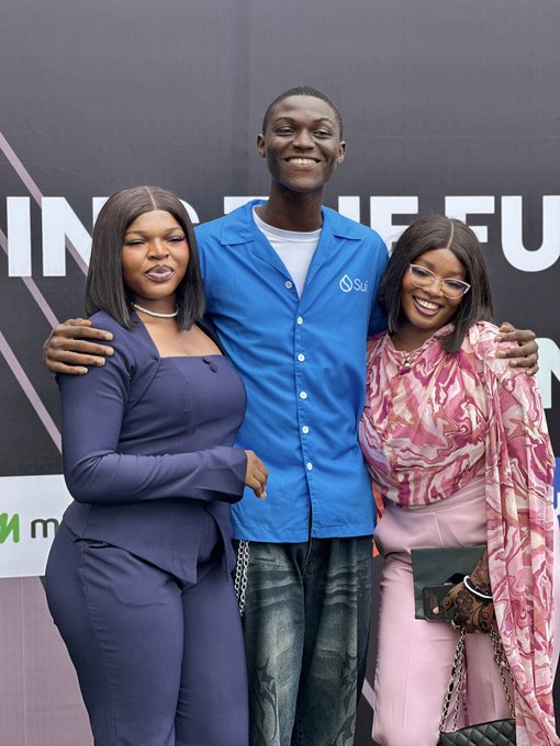 Three people pose together at an event: two women flank a tall man wearing a blue Sui-branded shirt standing in front of a backdrop with partial text '...HE FU...'.