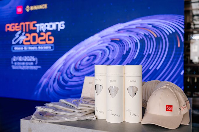 A promotional display for 'AGENTIC TRADING Era 2026' by Binance, featuring stacked 'ROUTINE' tumblers, a beige 'SSI' cap, and packaged merchandise on a counter with a large digital screen backdrop.