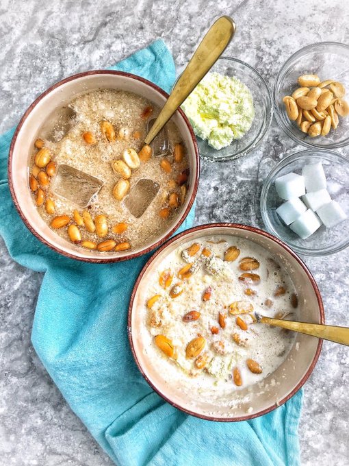 Two bowls of a creamy beverage topped with peanuts and ice cubes are arranged with small dishes of peanuts, lime zest, and sugar cubes on a teal cloth.