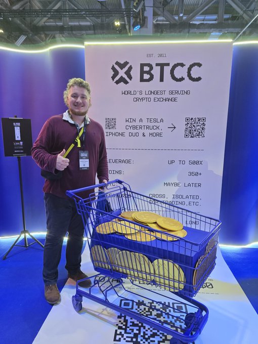 A man in a maroon sweater poses at a BTCC crypto exchange booth with a shopping cart full of gold BTCC coins; the banner reads 'EST. 2011', 'WORLD'S LONGEST SERVING CRYPTO EXCHANGE', 'LEVERAGE: UP TO 500X', 'COINS: 350+', and lists features like HAYBE LATER, CROSS, ISOLATED, and PORTFOLIO ETC.