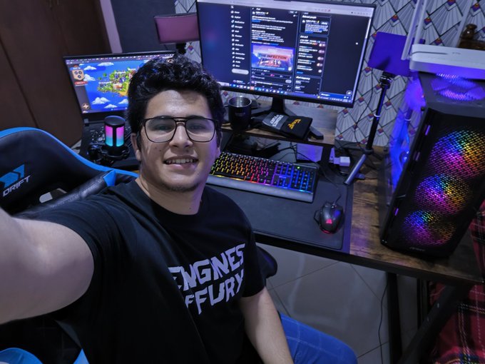 A young man with glasses smiles for a selfie at his gaming setup, which features dual monitors, an RGB keyboard, and a gaming PC with RGB lighting.