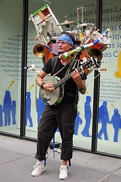 A street performer playing a banjo while wearing an elaborate costume covered in various electronic devices and computer hardware components.