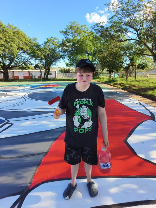 A young boy stands on an outdoor basketball court holding a water bottle and an orange popsicle, wearing a black shirt that reads 'MY FAVOURITE PEOPLE CALL ME POPS' with a cartoon illustration.