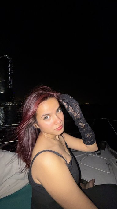 A woman with reddish-purple hair wearing a black top and black lace gloves posing at night on what appears to be a boat with a city skyline in the background.