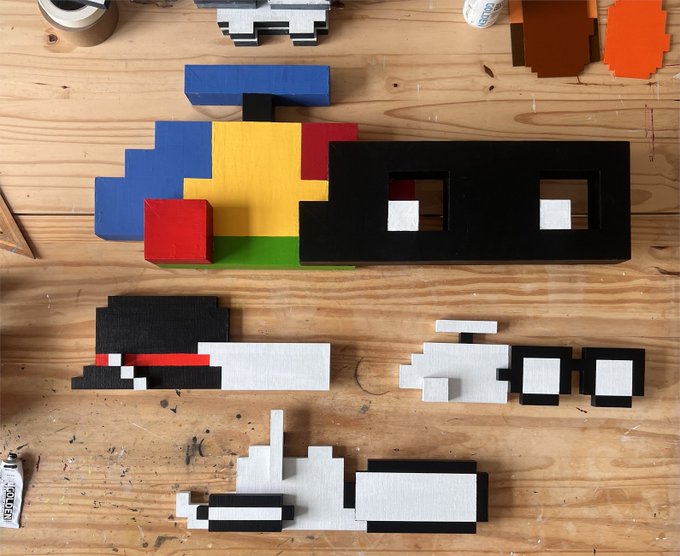 An assortment of colorful pixel art pieces and blocks made from materials like wood and foam, including primary colors and black and white shapes, laid out on a wooden workbench.