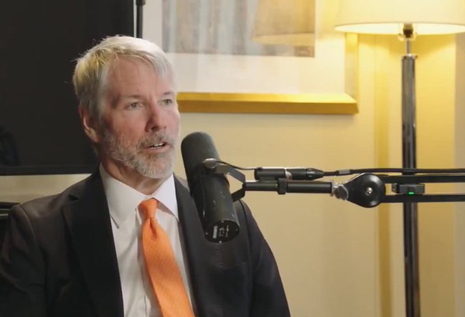 Michael Saylor, wearing a dark suit and orange tie, speaks into a podcast microphone. The image accompanies his bold prediction that Bitcoin will surpass Gold's market cap by 2035.