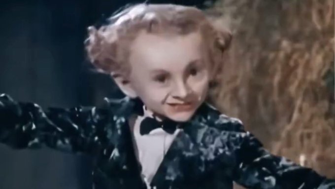 A vintage film still showing a young boy with curly blonde hair wearing a dark, shiny jacket and bow tie, smiling playfully.