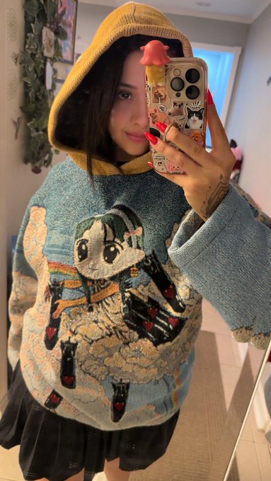 A mirror selfie of a person wearing a yellow hood and a colorful sweater featuring a Stitch character design, holding a phone with decorative charms.