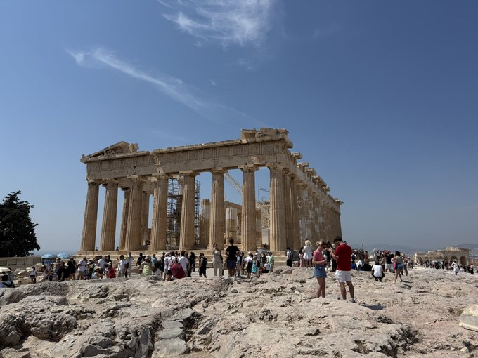 Tourists gather around the ancient Parthenon on the rocky hilltop of the Acropolis in Athens under a clear blue sky.