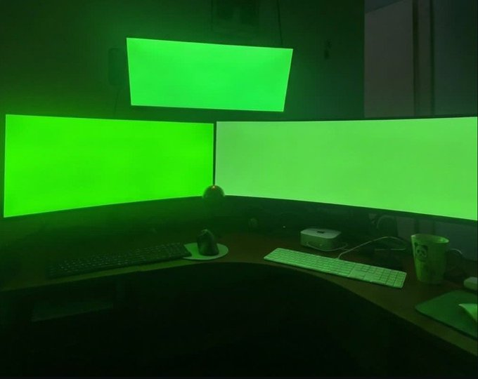 A dark room featuring three computer monitors displaying solid green screens, set up on a desk with a keyboard, mouse, and mug.