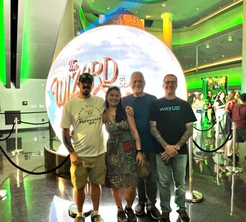 Four people pose together in front of a large circular display featuring "The Wizard of Oz" promotional artwork in what appears to be a cinema lobby.