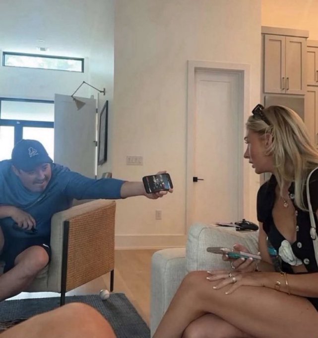 A man in a blue cap and shirt showing his phone to a blonde woman in a living room setting, with the woman appearing focused on her own phone.