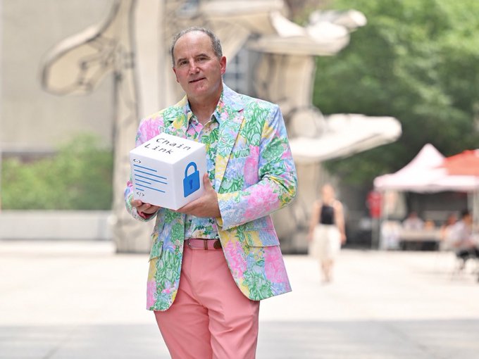 A smiling man in a colorful pink and green floral blazer holds a white box labeled "ChainLink" while standing outdoors near a sculpture, appearing to attend the Chainlink LINK:NYC event mentioned in the post.