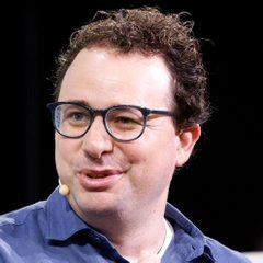A curly-haired man wearing blue-rimmed glasses and a denim shirt speaks into a lapel microphone, appearing to be at a public speaking event. The image likely depicts an Anthropic executive, possibly CEO Dario Amodei, discussing the company's reported $42B net loss despite 12x revenue growth and a potential $2T IPO valuation.