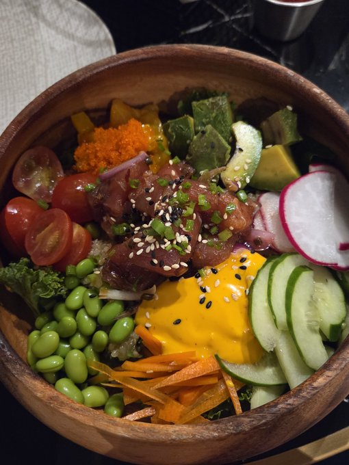 A wooden bowl filled with a colorful poke-style dish containing tuna, avocado, cucumber, edamame, carrots, cherry tomatoes, radish, mango, tobiko, and sesame seeds over rice.