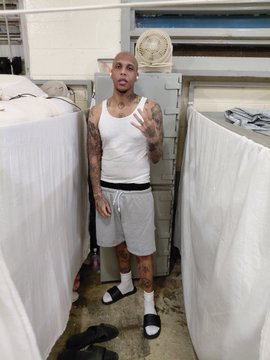 A heavily tattooed man in a white tank top and gray shorts stands in what appears to be a laundry room, making a hand gesture toward the camera. The post references onboarding him onto "Fomo" (likely a social platform), suggesting the man's distinctive look made an impression worth pursuing.