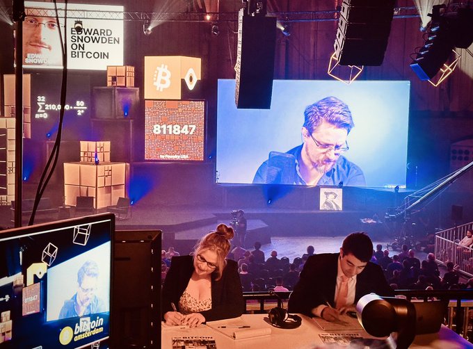 At the Bitcoin Amsterdam 2023 conference, two broadcasters sit at the "Livedesk" with Edward Snowden appearing on the large screen behind them, beneath banners reading "Edward Snowden On Bitcoin" and displaying Bitcoin's price at $29,210. The scene captures the surreal moment when Snowden's talk about freedom and Bitcoin unfolded live at the event.