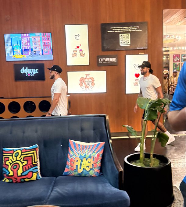 Two men in white t-shirts and black caps walk through a vibrant art gallery with framed cartoon-style illustrations on wood-paneled walls, while a blue tufted couch with colorful 'SPLASH' and Keith Haring-style pillows sits in the foreground next to a potted plant.