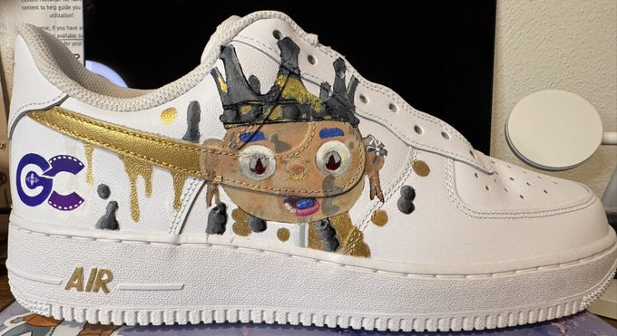 A customized white sneaker featuring hand-painted artwork including a character with a crown, purple 'GG' letters, gold paint drips, and 'AIR' branding on the sole.
