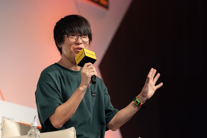 A man wearing glasses and a dark green t-shirt speaks into a yellow 'Block' branded microphone while gesturing with his other hand, with a colorful wristband visible.