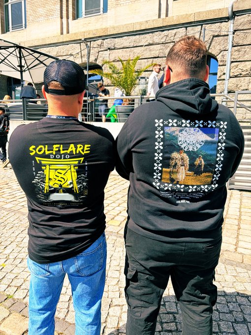 Two people stand outdoors on a cobblestone surface wearing black t-shirts, one with a yellow "SOLFLARE DOJO" graphic and the other with a large artistic design on the back.