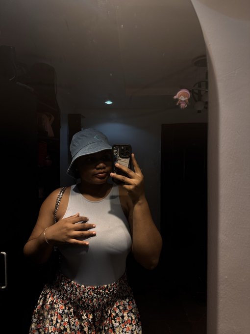 A person wearing a denim bucket hat, white sleeveless top, and floral skirt takes a mirror selfie with their phone in a dimly lit room.