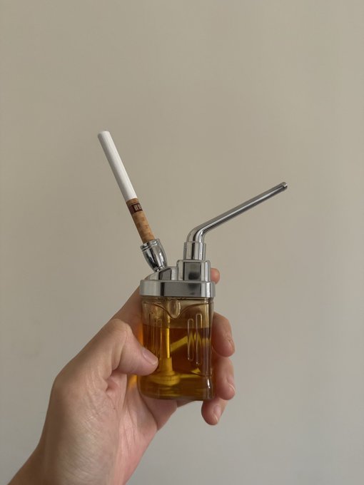 A hand holds a small, clear glass vape or device filled with amber-colored liquid, featuring a chrome top, a metal mouthpiece, and an attached cigarette-style tip.