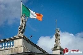 An Irish flag waves in the sky above ornate stone statues on a building's rooftop, with a 'DinheiSK ON OFACADA' watermark in the corner.