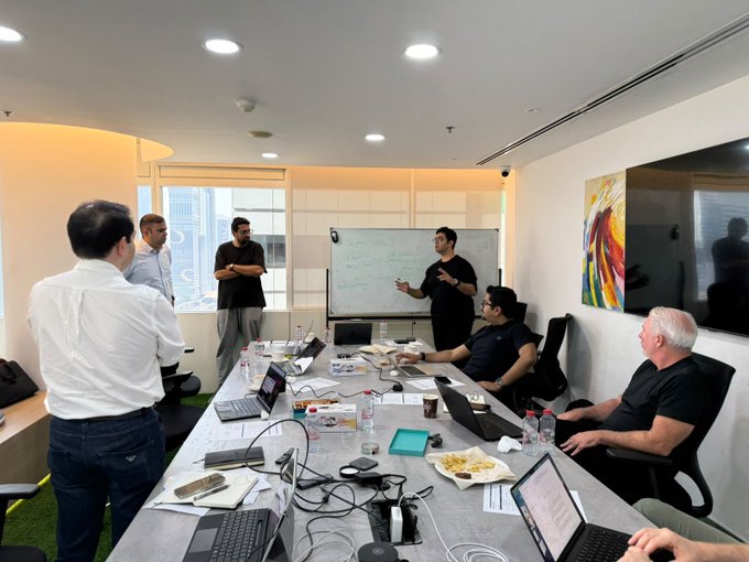A group of professionals gather around a conference table covered with laptops, cables, and a plate of food, while a presenter in a black shirt gestures in front of a whiteboard in an active office meeting. The whiteboard session ties into the post's announcement of a major "cook" event for $ZIG, hosted in partnership with Disrupt.com and Zamanat, suggesting this is a strategic planning or development meeting for the collaboration.