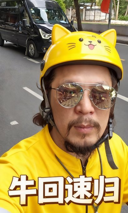 A man wearing a yellow cat-eared helmet and reflective aviator sunglasses poses for a selfie on a street, wearing a yellow shirt with the Chinese text '牛回速归' overlaid on the image.