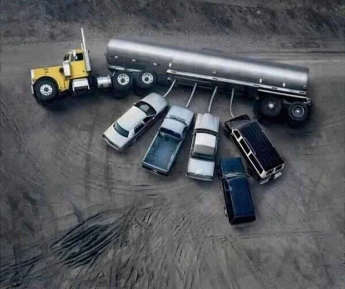 An aerial photograph of a yellow tanker truck with multiple hoses connected to several cars parked in a dirt lot.