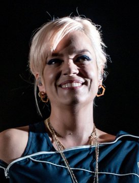 The image shows a smiling blonde woman in a dark blue outfit with layered gold necklaces and gold hoop earrings, appearing to be at a performance or event. The post references Lily Allen, suggesting this is the singer pictured, in connection with the story of her missed opportunity to receive Bitcoin worth $17 billion today for a 2009 Second Life streaming gig.