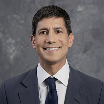 A smiling man in a dark suit, white shirt, and blue tie poses against a gray studio backdrop, likely a financial analyst or expert providing commentary on the Federal Reserve's 25 basis point interest rate hike and its impact on Bitcoin ($BTC) and Ether ($ETH).