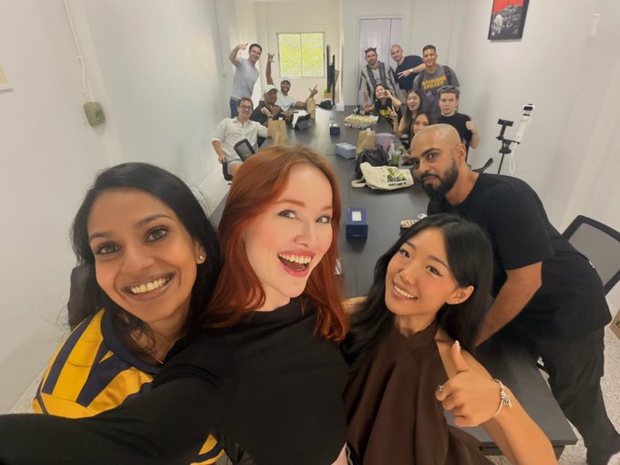 A group selfie of smiling people gathered around a long table in a meeting room, with three women in the foreground and several others seated further down the table.