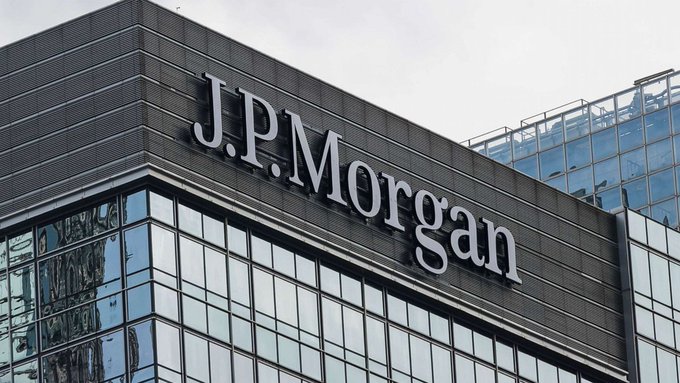 The image shows a large building with J.P. Morgan written on its side, located under a cloudy sky. The words "J.P. Morgan" are clearly visible. In addition to the building, there are various windows that reflect light throughout the scene. One of these windows has the words "CRAZY: $10,000,000,000+" written in bold letters on it, which is related to a recent news story about cryptocurrency trading happening at J.P. Morgan. The post also mentions other cryptocurrency details and a tokenized U.S. Treasury settlement involving the $XRP Ledger, further highlighting the connection between the building and the cryptocurrency market.