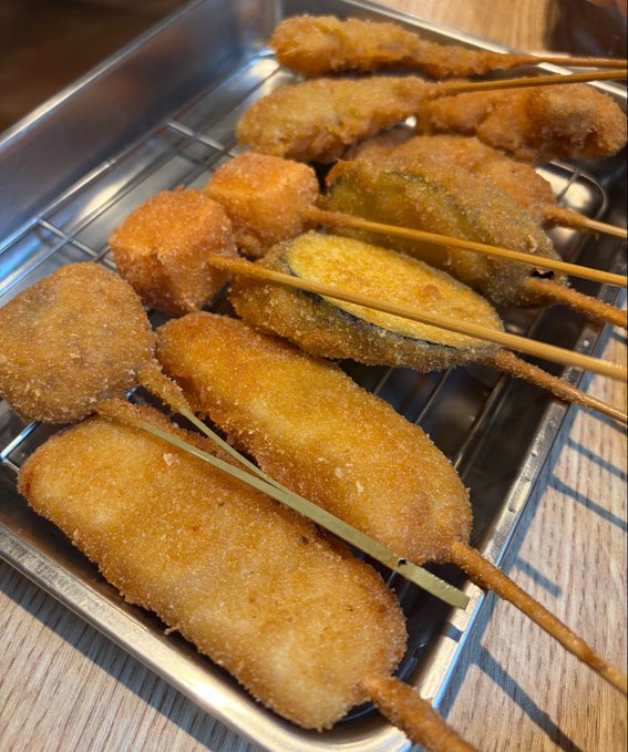 A close-up photo of Japanese kushikatsu (deep-fried skewers) served on a metal grill tray, including various breaded items on bamboo sticks.