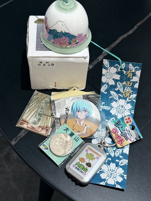 A collection of Japanese souvenirs arranged on a black table, including a Mt. Fuji wind chime resting on its box, a "大雪山" (Daisetsuzan) commemorative coin, an anime character badge, a San-X acrylic charm (appears to feature characters like the bear), a Yoshida Paper Company bookmark tagged "有田焼" (Arita-yaki), and other small trinkets. The image reflects the caption's sentiment of a delighted recipient showing off thoughtful gifts brought back from Japan.
