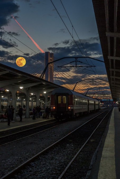 The image shows a train station with people waiting for their trains at night. The sky has a beautiful sunset, creating a serene atmosphere. A full moon can be seen in the background, adding to the overall ambiance of the scene. The photo is taken from an elevated perspective, providing a clear view of the train tracks and the surrounding area.
