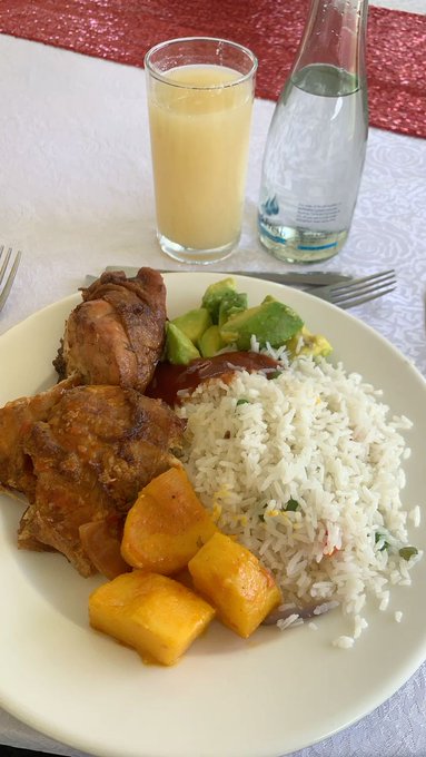 The image shows a plate of food consisting of rice, chicken, and vegetables such as avocado and squash. A glass of orange juice is also present on the table next to the plate. The scene appears to be set for breakfast or brunch with a variety of dishes served together.