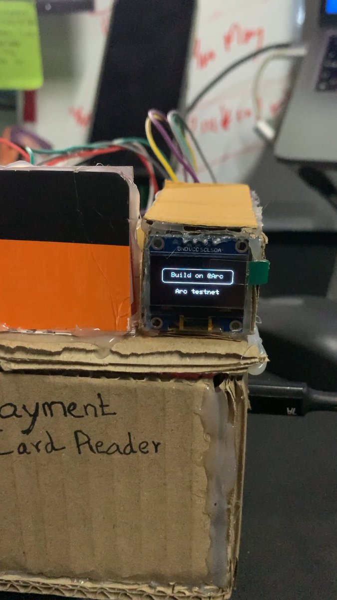 A cardboard DIY payment card reader prototype featuring a small OLED screen displaying "Build on @Arc" and "Arc testnet," alongside jumper wires and electronic components—reflecting the hackathon project teased in the post.